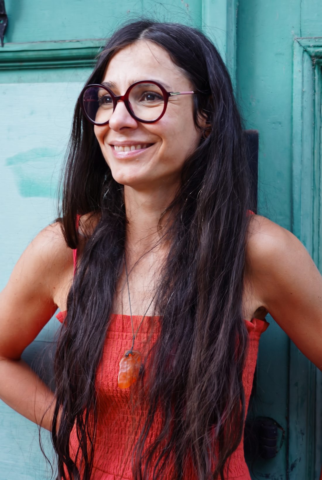 Deniza Miteva mit runder Brille und terracottafarbenem Trägertop, Kette mit Bernstein-Anhänger, der Blick zur Seite, vor einer türkisen Holztür